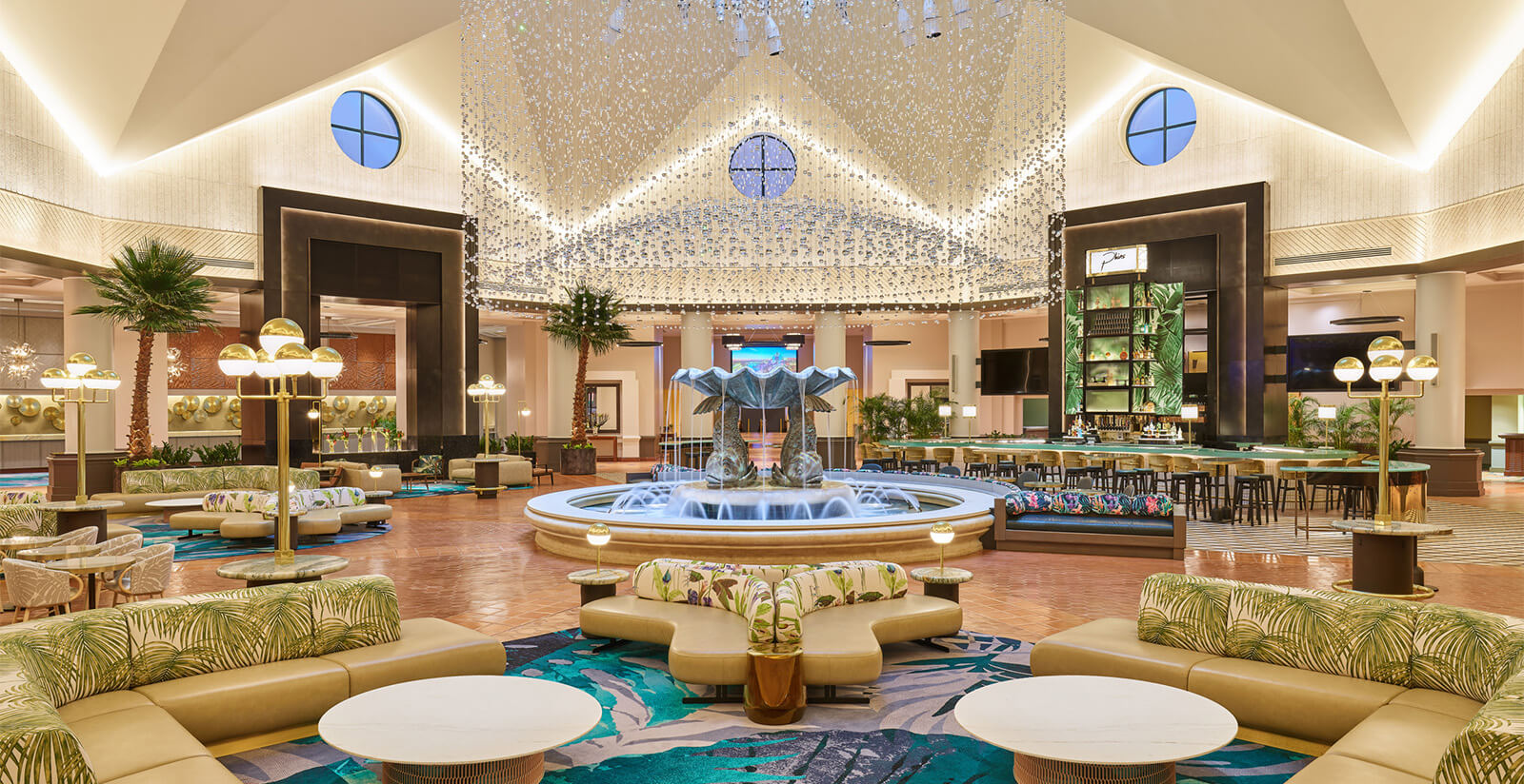 Spacious hotel lobby with a central water fountain, modern seating areas, decorative plants, and high ceilings with hanging lights; large windows and a stylish bar are visible in the background.