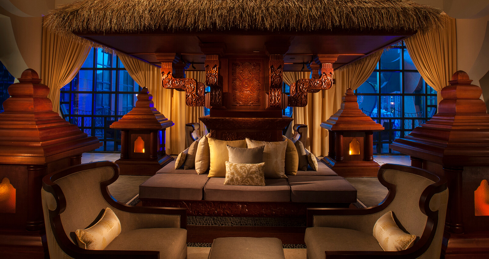 Luxurious, dimly lit lounge with carved wooden decor, thatched roof, cushioned seating, ornate lanterns, and draped curtains, creating an elegant, tropical ambiance. Large windows in the background reveal a blue-lit view.