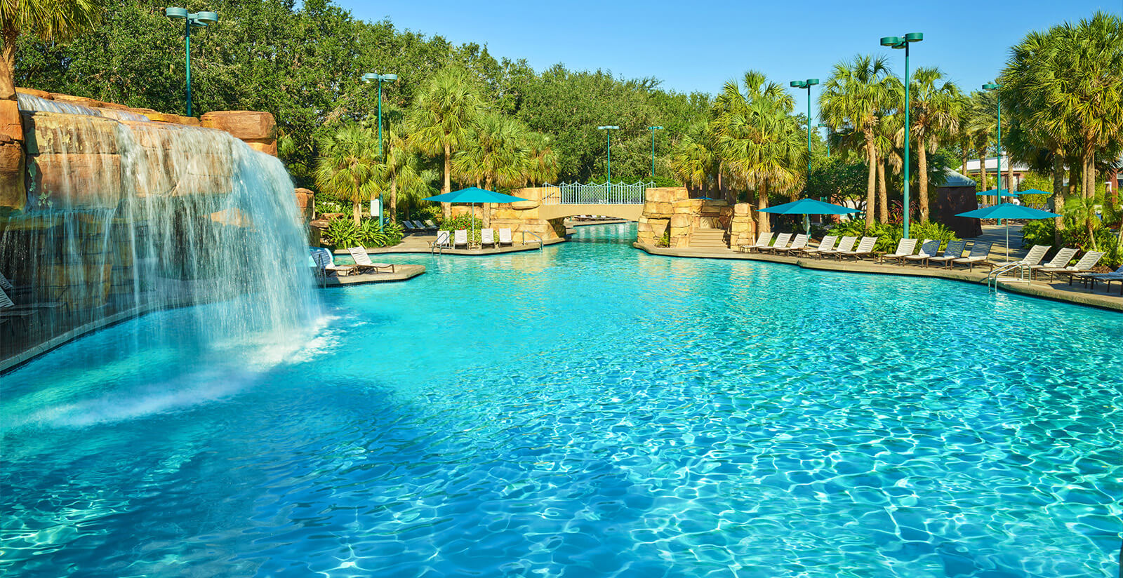 A large outdoor swimming pool with clear blue water features a waterfall on the left, lounge chairs, umbrellas, palm trees, and lush greenery under a clear blue sky.