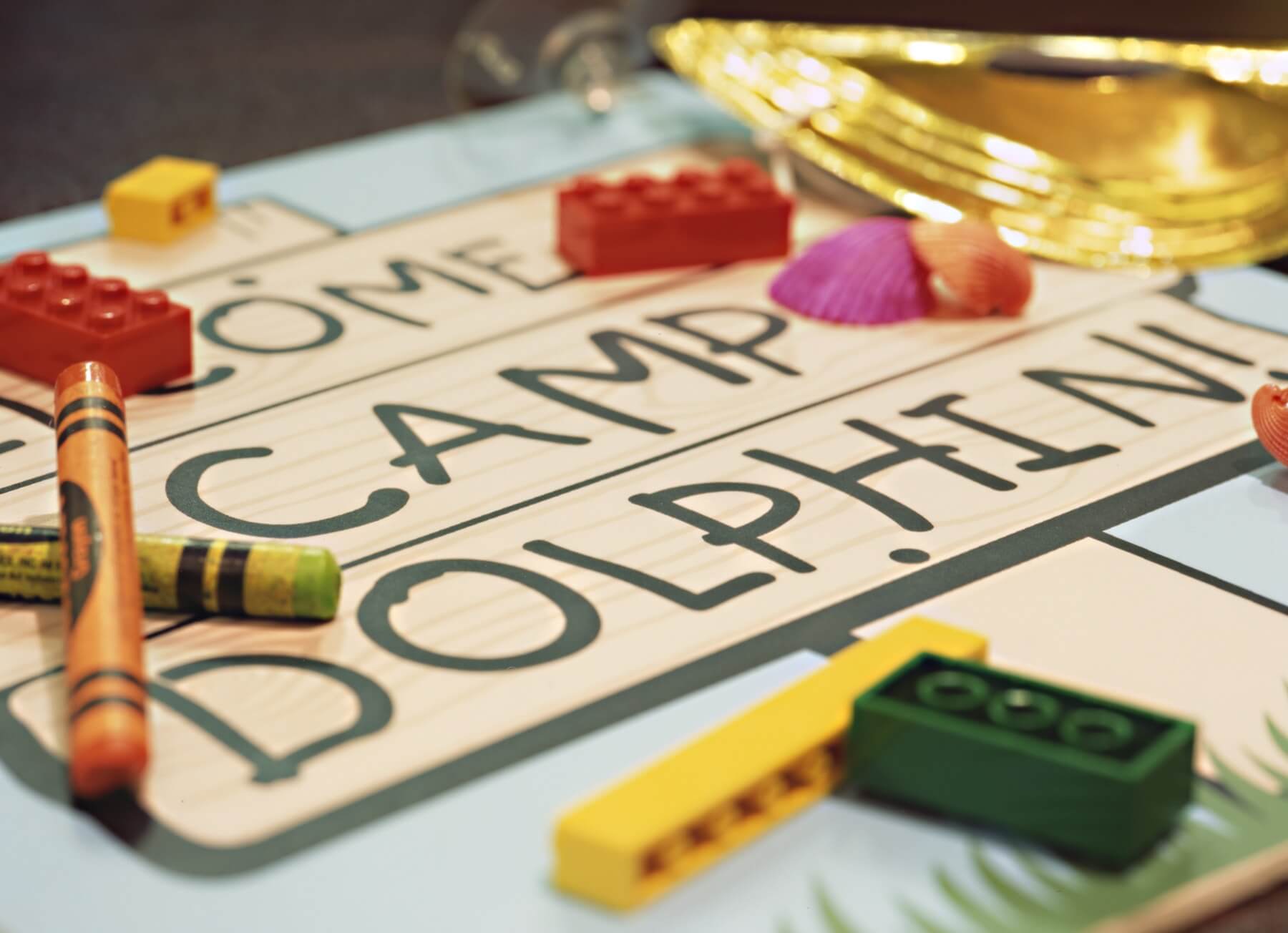 Close-up of a sign reading “CAMP DOLPHIN” with crayons, colorful toy building blocks, a seashell, and part of a shiny gold party hat scattered on top.