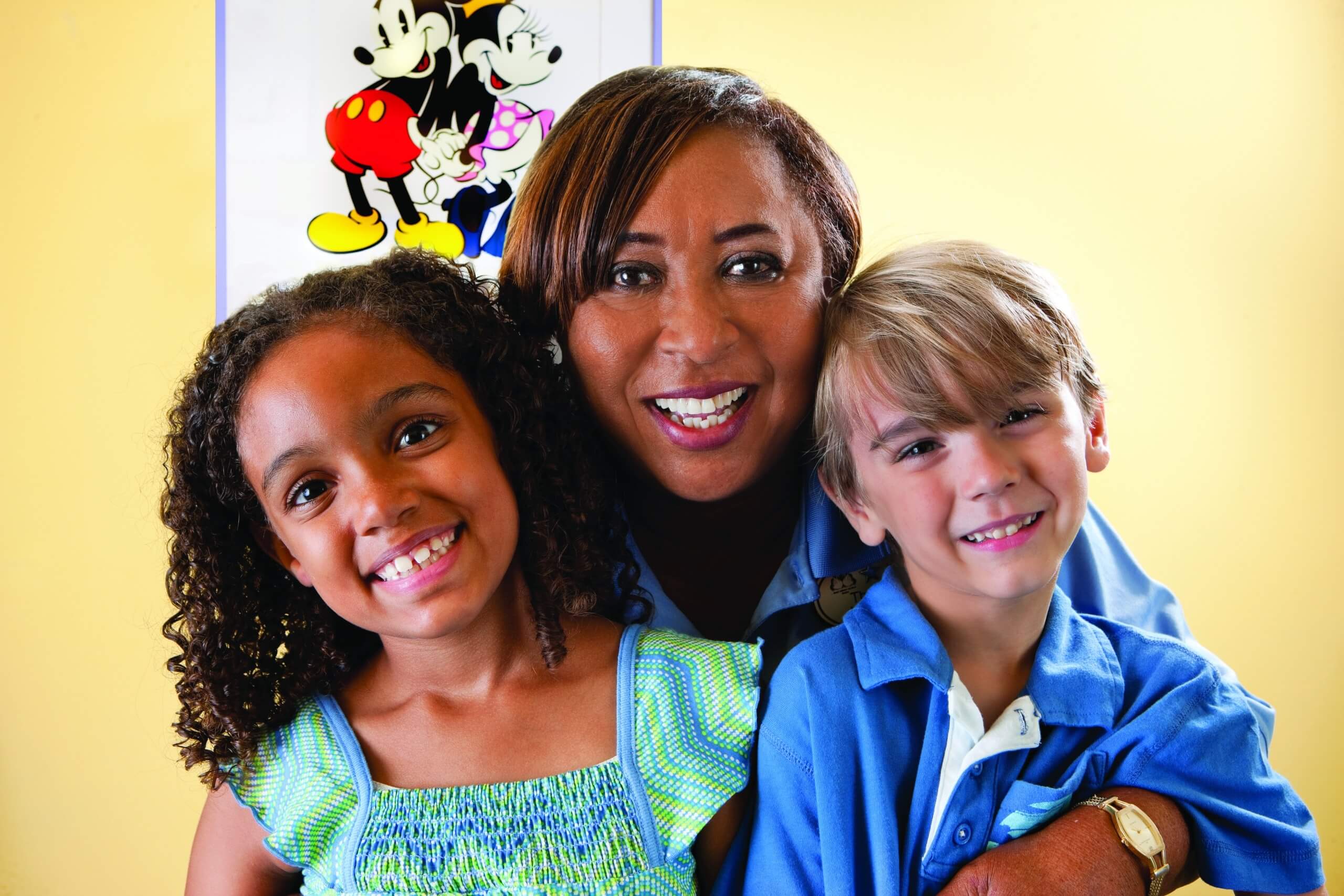 A woman smiles and hugs two young children, one girl and one boy, in front of a Mickey and Minnie Mouse poster on a yellow wall. All three look happy and cheerful.