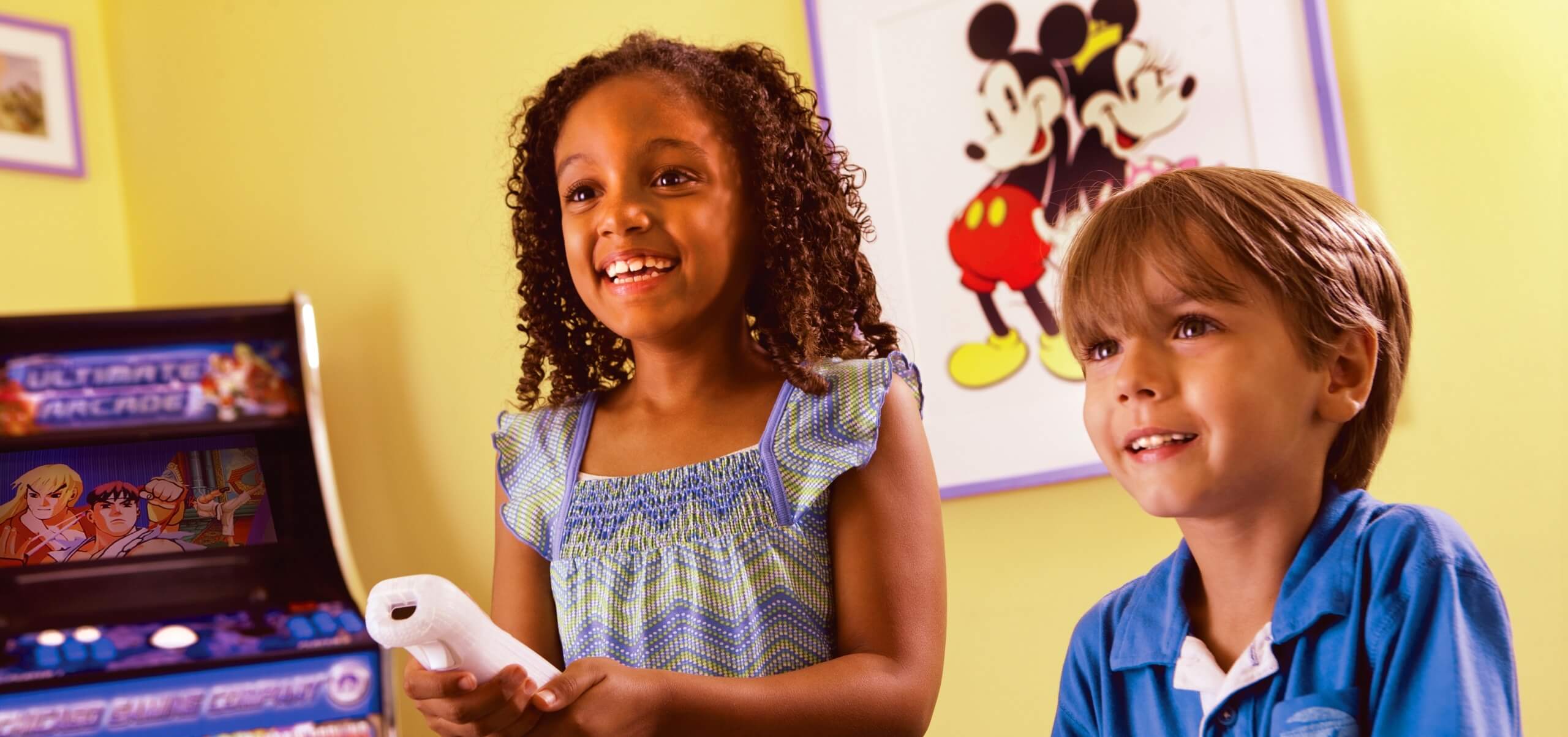 Two children, a girl holding a game controller and a boy in a blue shirt, smile while playing a video game. Behind them is a framed picture of Mickey and Minnie Mouse on a yellow wall and an arcade machine to the left.