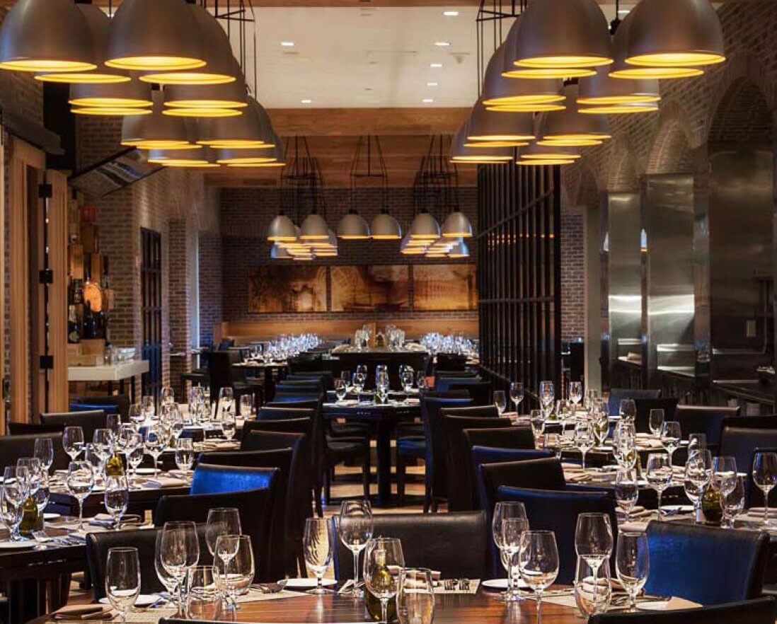 A modern, elegant restaurant with brick walls, wooden tables, dark chairs, and hanging pendant lights. Tables are set with glassware and silverware, creating a warm, inviting atmosphere.