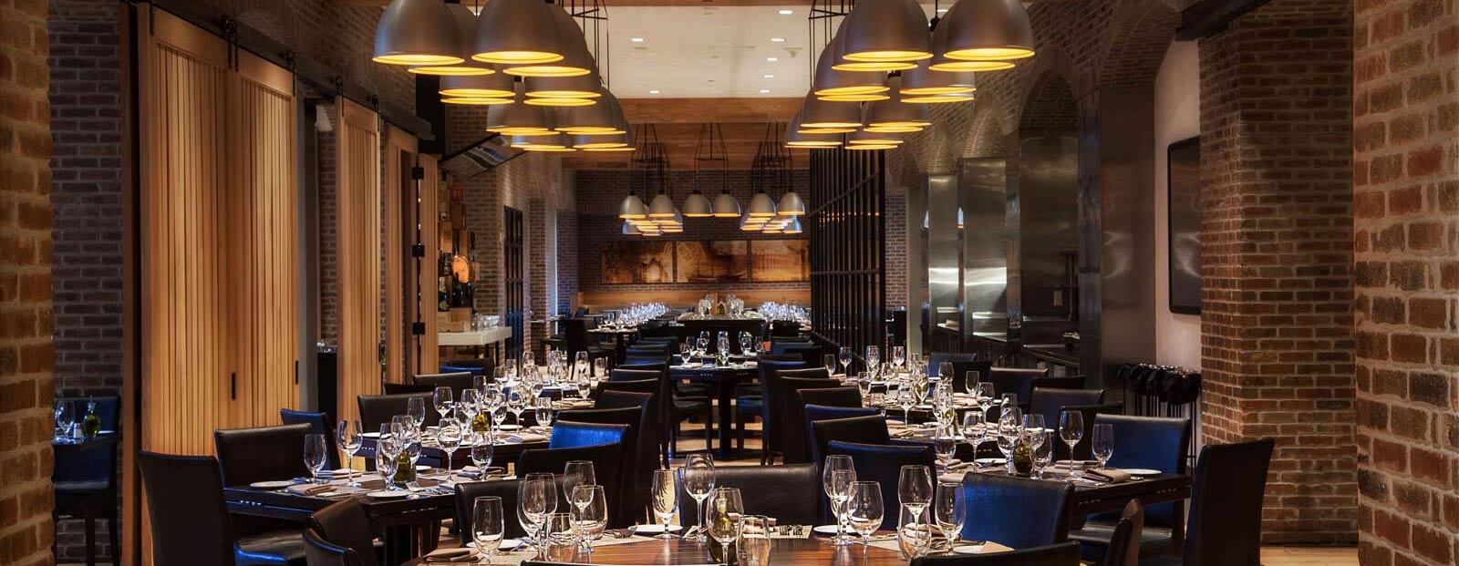 A modern, elegant restaurant with brick walls, wooden tables, dark chairs, and hanging pendant lights. Tables are set with glassware and silverware, creating a warm, inviting atmosphere.