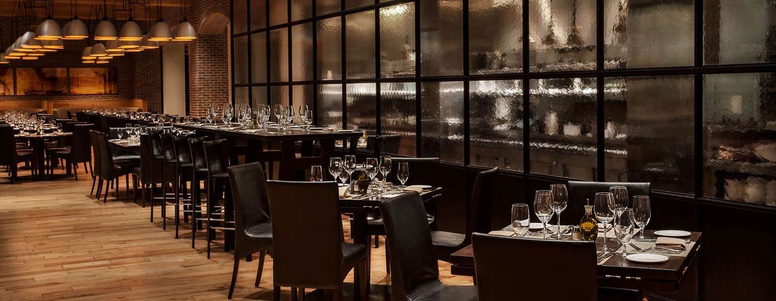 A modern, upscale restaurant with dark wooden tables and chairs, set with glassware and silverware. Warm lighting hangs over the dining area, and a glass partition separates the spaces. The atmosphere is elegant and inviting.