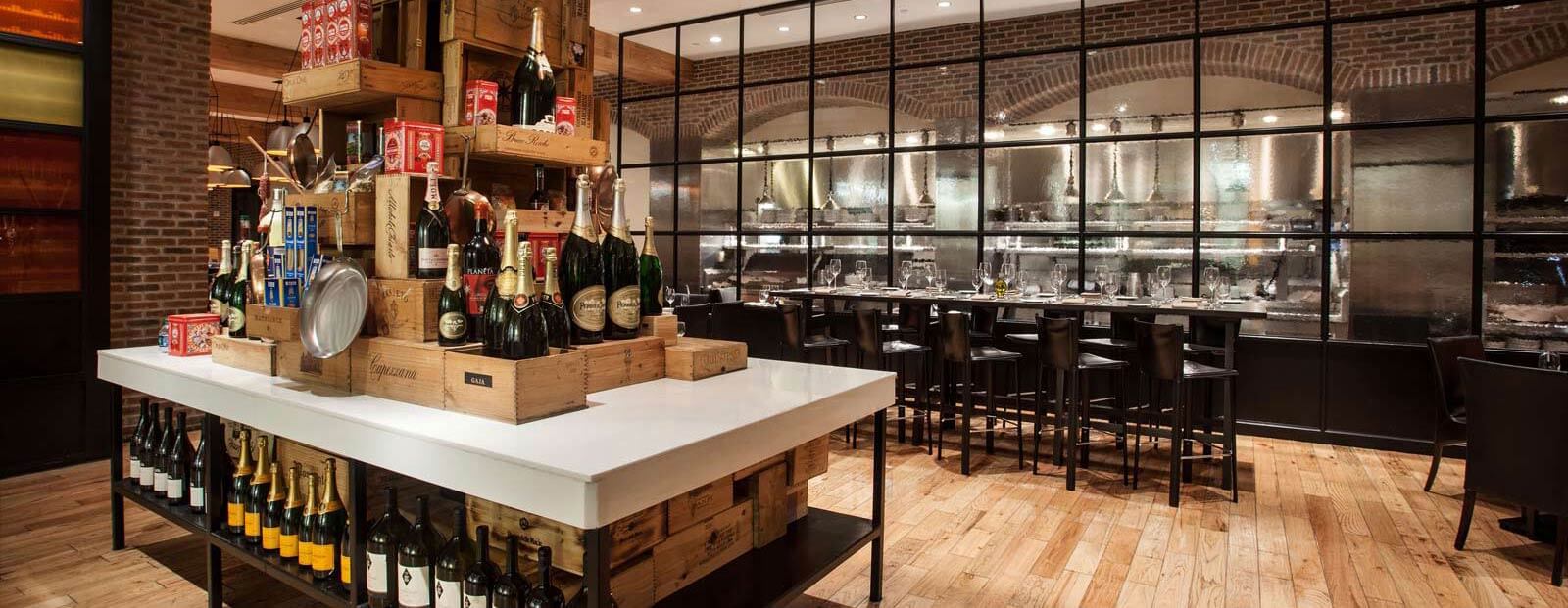 A modern restaurant interior with a wooden floor, a large island displaying wine bottles and kitchen items, and a dining area with black chairs and tables by a glass wall separating the kitchen.