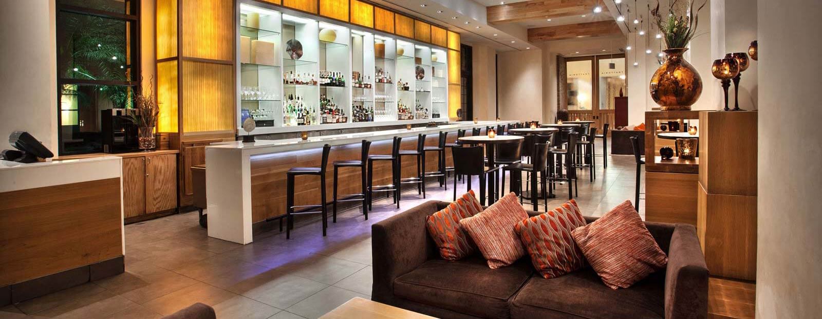 A modern, stylish bar with a long counter, high stools, backlit shelves stocked with bottles, a brown sofa with patterned cushions in the foreground, and warm ambient lighting throughout the space.