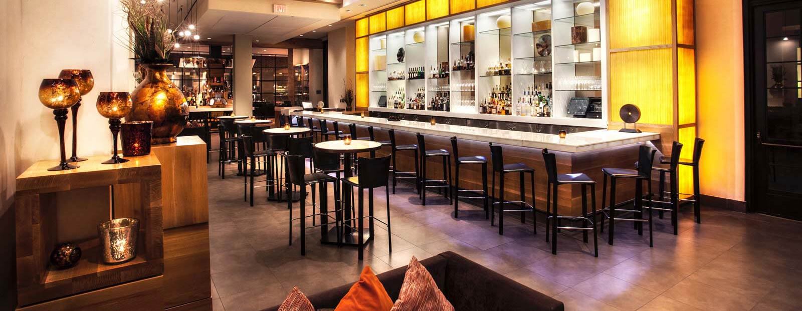Modern bar interior with a long white marble counter, high black chairs, illuminated shelves with bottles, small round tables with chairs, warm lighting, and decorative vases, creating an inviting atmosphere.