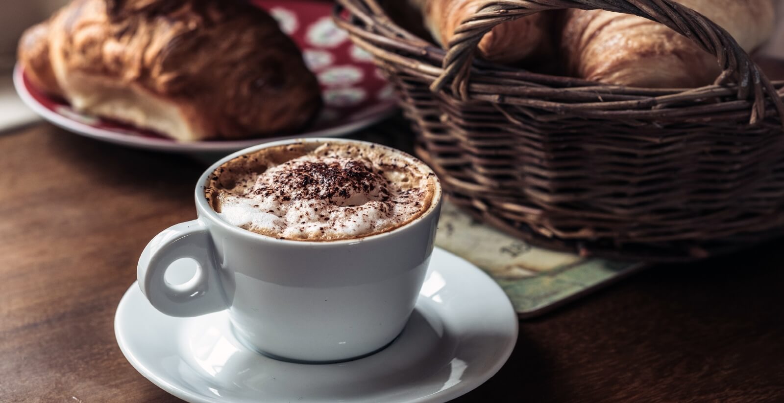 A white cup of cappuccino with frothy milk and chocolate sprinkles sits on a saucer beside a wicker basket filled with croissants and a plate of pastries on a wooden table.