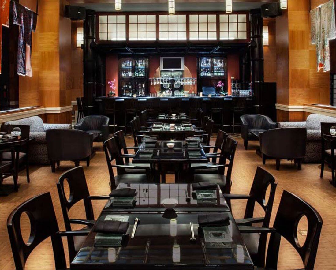 Elegant restaurant interior with dark wooden tables and chairs neatly arranged, soft lighting, and a bar at the back. Colorful fabric panels hang on the walls, creating a warm and inviting atmosphere.