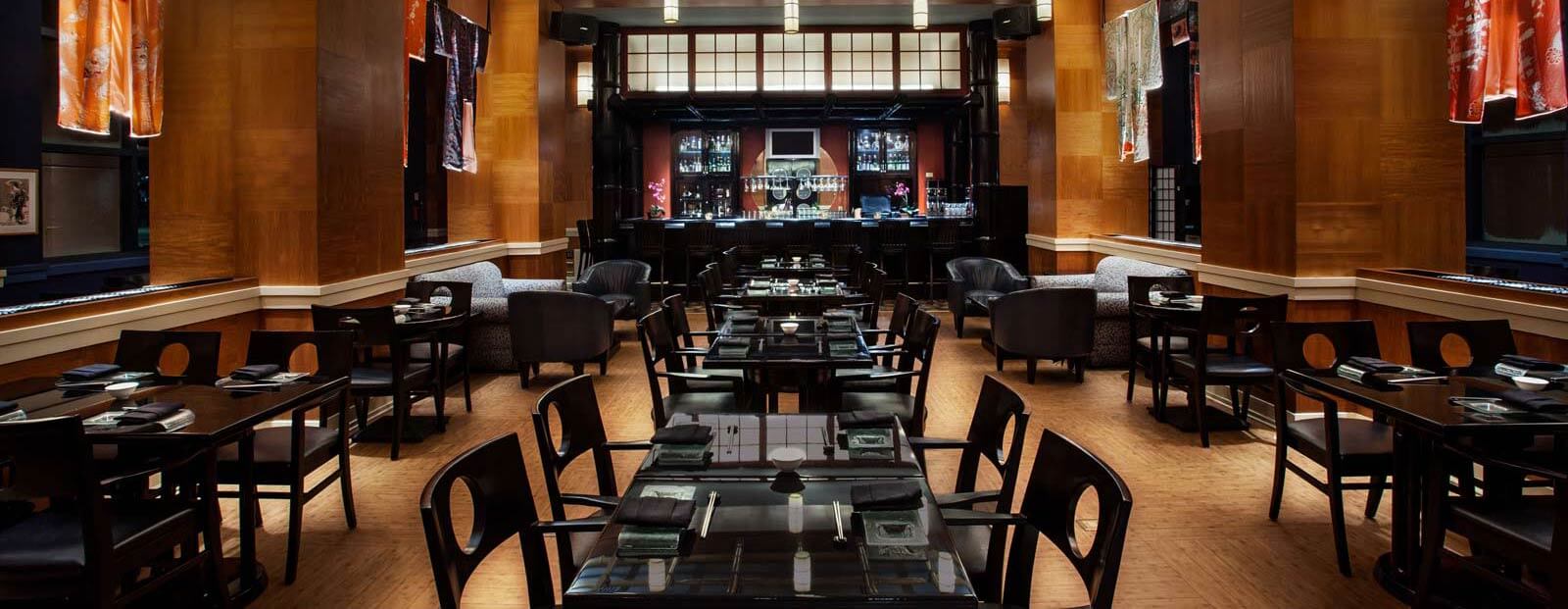 Elegant restaurant interior with dark wooden tables and chairs neatly arranged, soft lighting, and a bar at the back. Colorful fabric panels hang on the walls, creating a warm and inviting atmosphere.