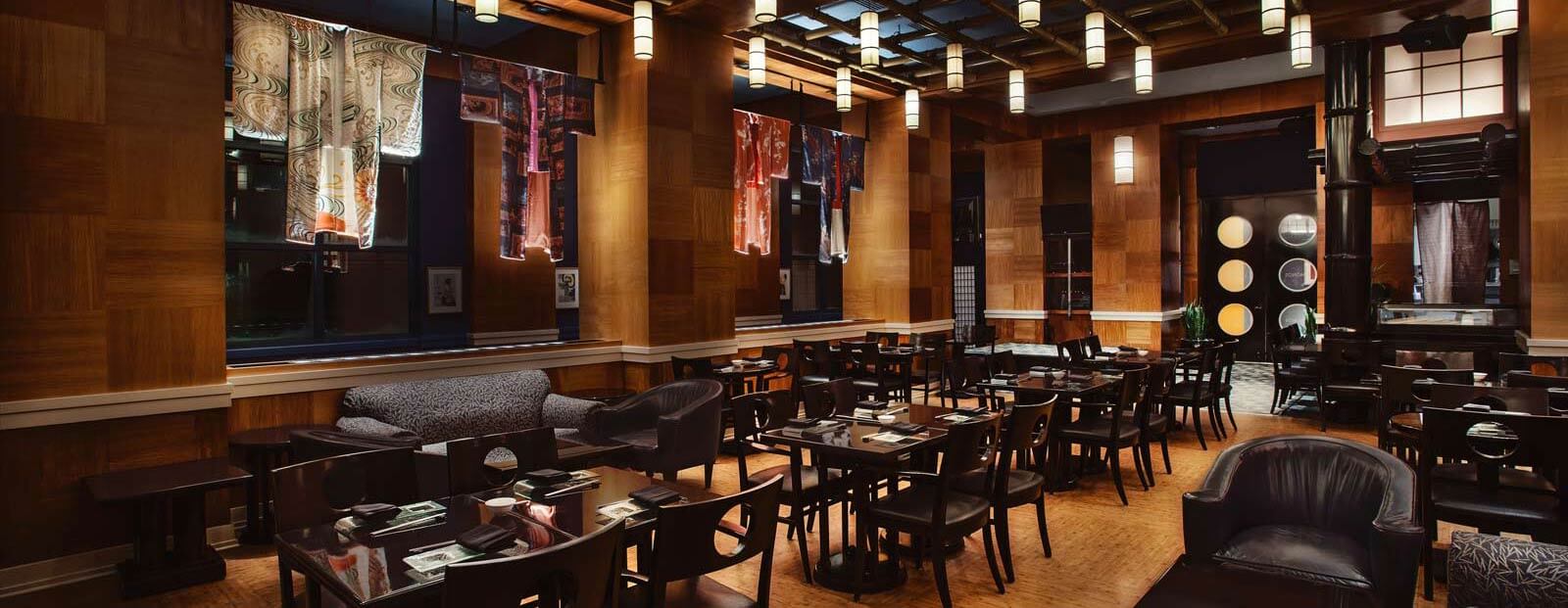 Elegant restaurant interior with wooden walls, modern hanging lights, dark chairs, and neatly set tables. Colorful fabric artwork is displayed in the windows, creating a sophisticated and inviting atmosphere.