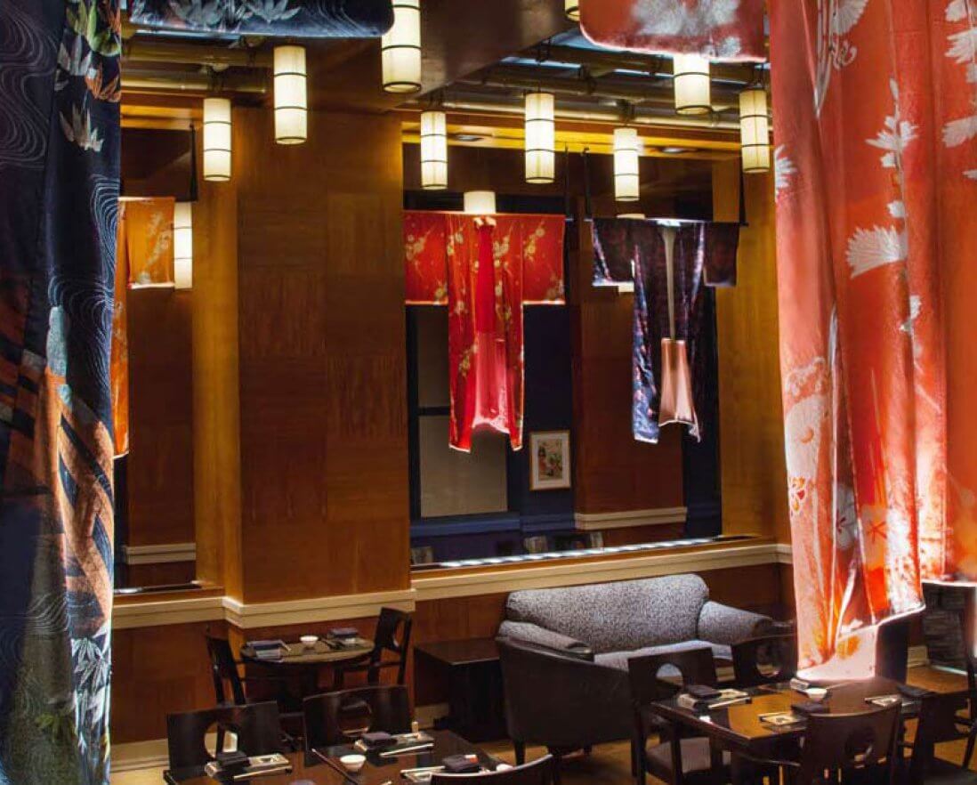 Colorful kimonos hang from the ceiling in a stylish restaurant with wooden walls, modern light fixtures, and tables set for dining, creating a warm and elegant atmosphere.
