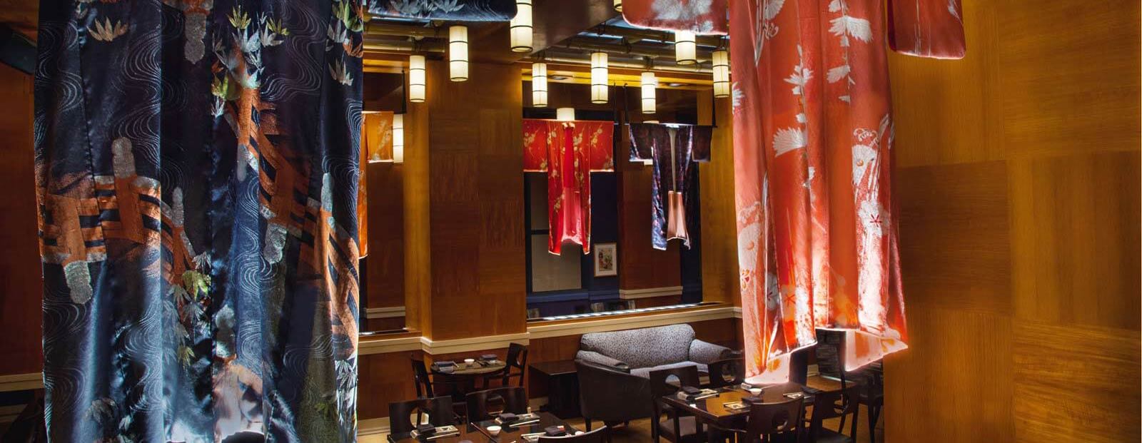 Colorful kimonos hang from the ceiling in a stylish restaurant with wooden walls, modern light fixtures, and tables set for dining, creating a warm and elegant atmosphere.
