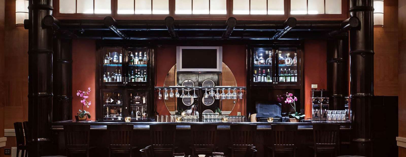 A stylish bar with dark wood furnishings, glass shelves holding bottles, hanging wine glasses, two pink orchid plants, and a large circular mirror behind the counter, all softly lit by overhead lights.