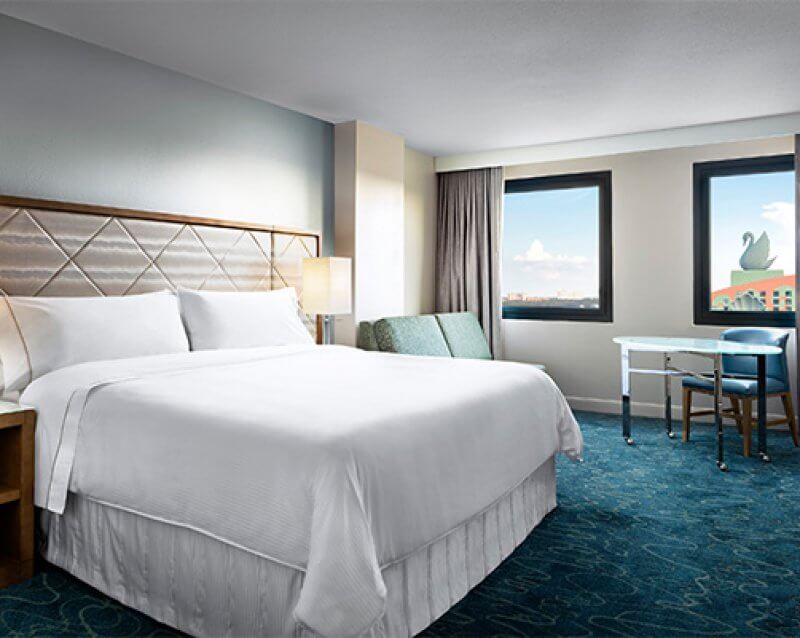 Ein modernes Hotelzimmer mit einem großen Bett, weißer Bettwäsche, Nachttischen mit Lampen, einer grünen Couch, einem Schreibtisch mit Stuhl und zwei Fenstern, die ein Gebäude mit einer großen Schwanenstatue zeigen.