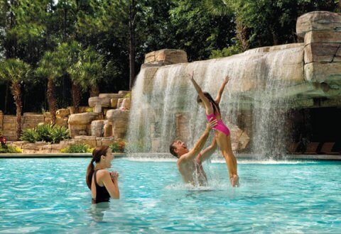 A man lifts a young girl in a pink swimsuit above a swimming pool, while a woman watches and smiles. Behind them is a large artificial waterfall and rock formation surrounded by palm trees.