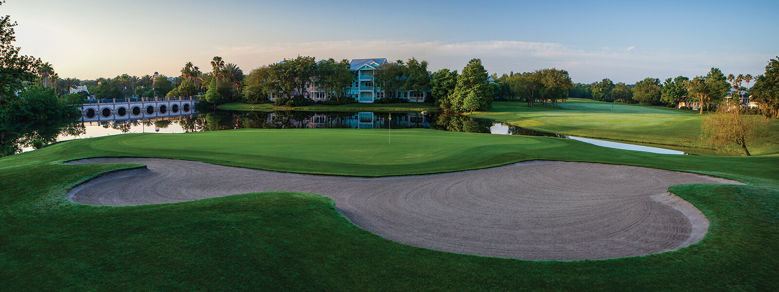 A panoramic view of a golf course featuring a large sand bunker in the foreground, a green with a flag, a calm water hazard, trees, a bridge, and buildings in the background under a clear sky.