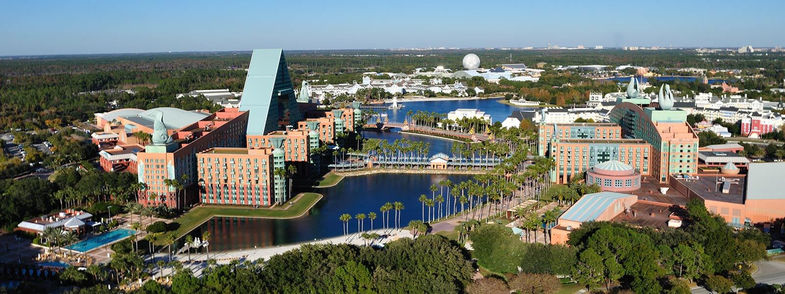 Aerial view of the Walt Disney World Swan and Dolphin Resort with lakes, pools, palm trees, and Epcot’s Spaceship Earth visible in the distance under a clear blue sky.