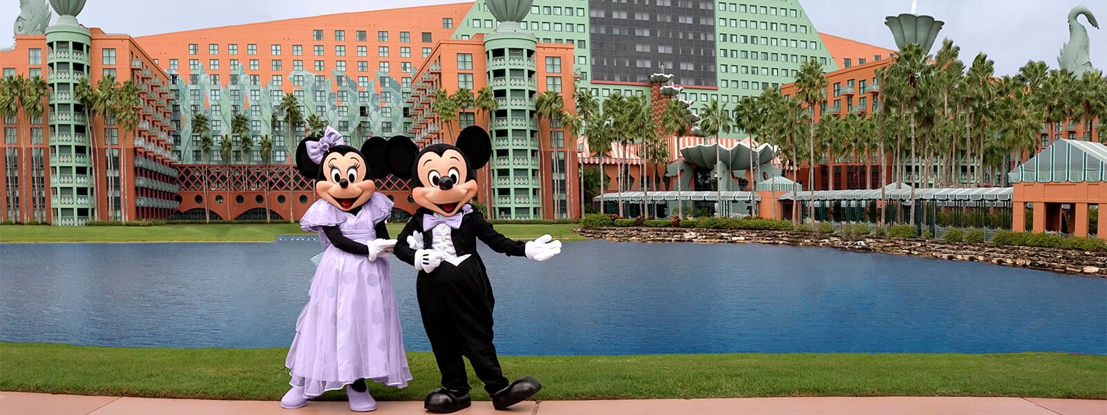 Minnie Mouse and Mickey Mouse, dressed in formal outfits, stand together smiling by a lake in front of the colorful Walt Disney World Dolphin Hotel, surrounded by palm trees.