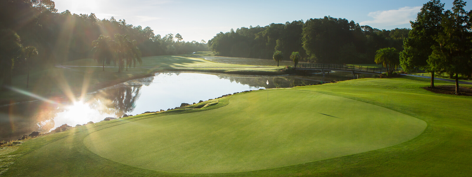 A scenic golf course at sunrise features a lush green putting area, a reflective pond, trees, and a wooden bridge under a clear blue sky, with sunlight streaming through the trees.