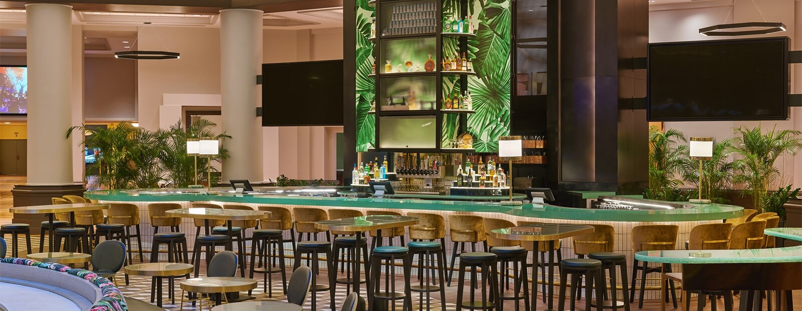 A modern hotel bar with a curved green countertop, tall bar stools, tropical leaf decor behind the shelves, and round tables with chairs, surrounded by potted plants and warm lighting.