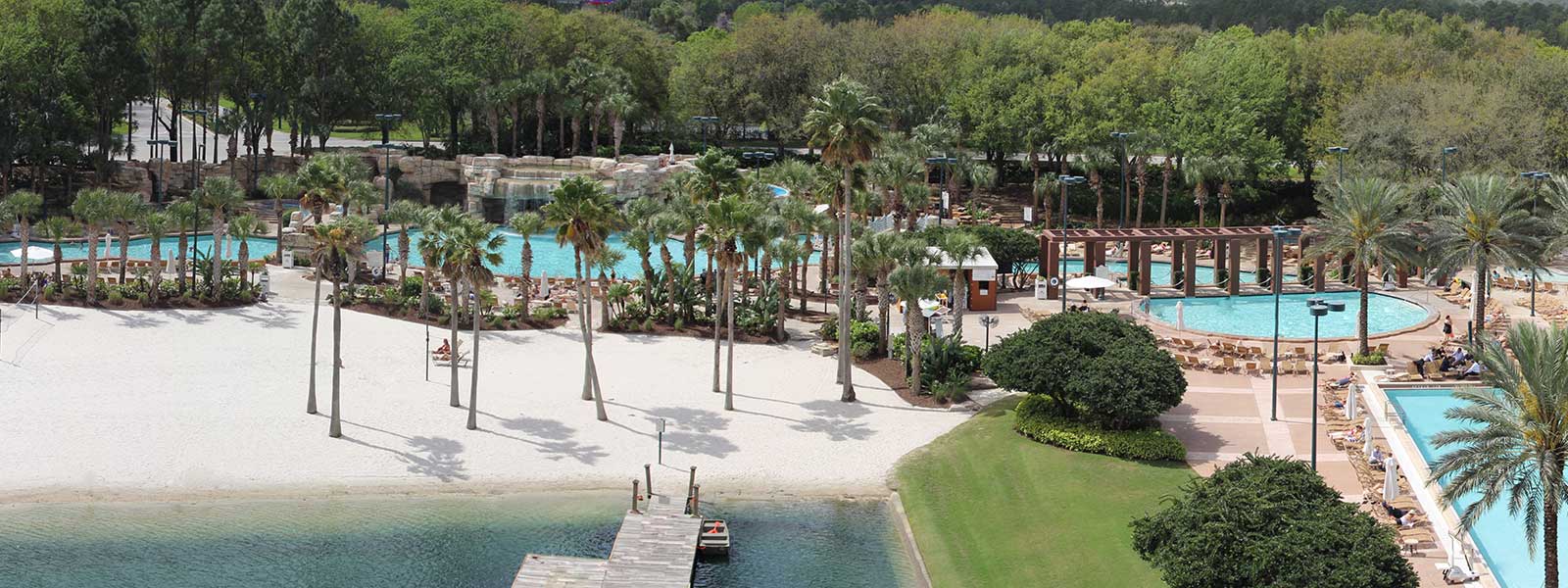 A resort pool area with palm trees, sandy beach, sun loungers, and a wooden dock extending into a lagoon, surrounded by lush greenery and forest in the background.
