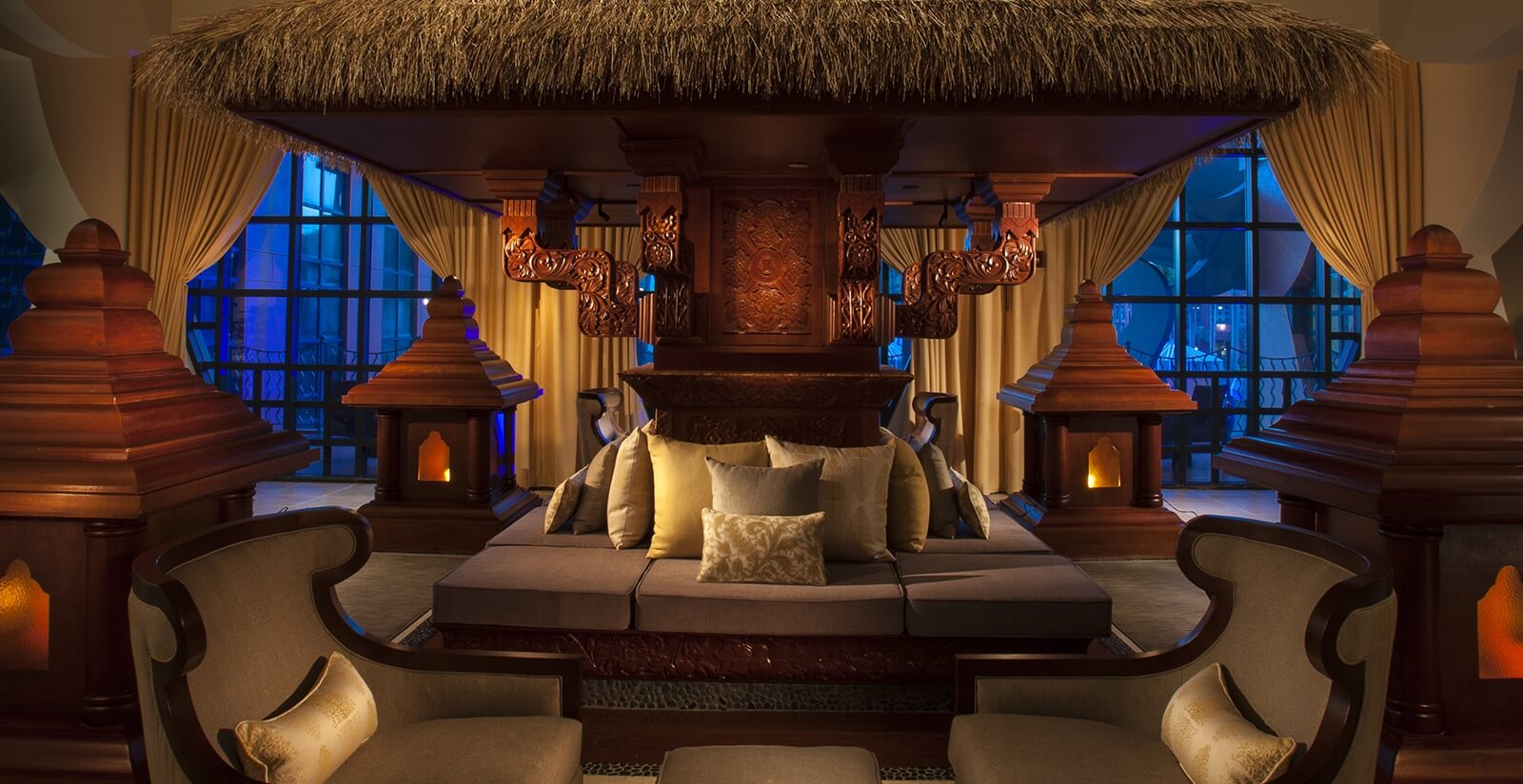 Luxurious lounge with ornate wooden decor, a thatched roof canopy, plush cushions, and ambient lighting, creating an inviting and elegant atmosphere with large windows in the background.
