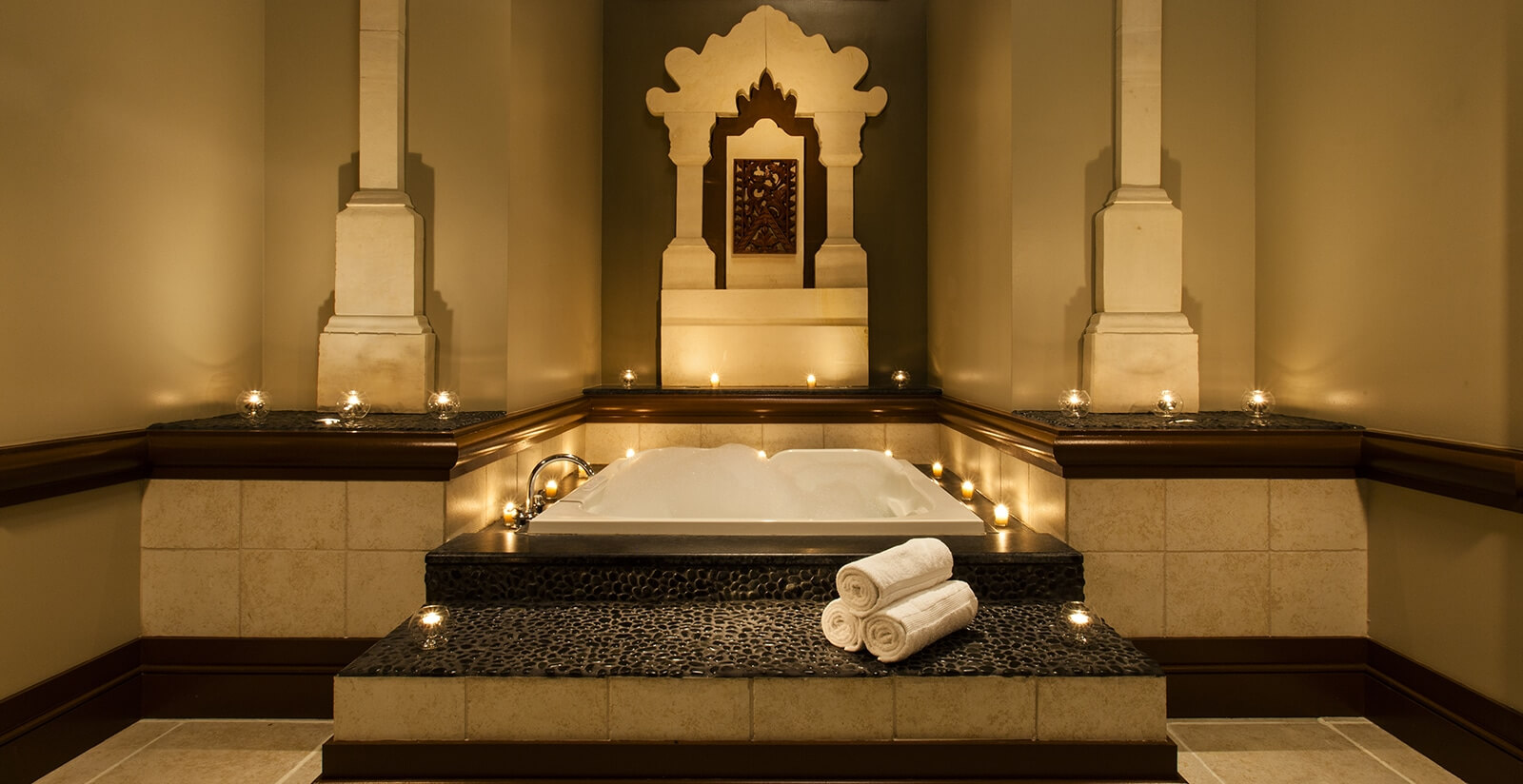 A luxurious spa bathtub is set in a decorative alcove with ornate architecture, surrounded by lit candles and rolled white towels, creating a serene and relaxing atmosphere.
