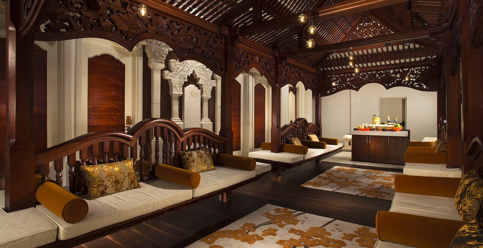 Luxurious wooden lounge with intricate carved details, cushioned seating along the walls with gold and brown pillows, warm lighting, and a refreshment table at the far end of the room.