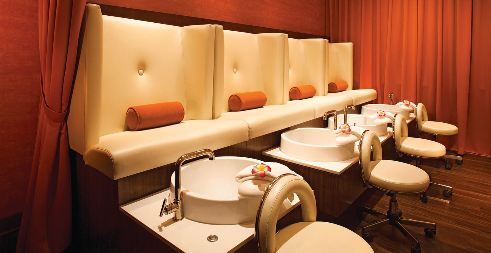 A luxurious spa pedicure area with cream-colored chairs, matching footrests, round pillows, white pedicure basins, and rolled towels with flowers, set against warm orange walls and curtains.