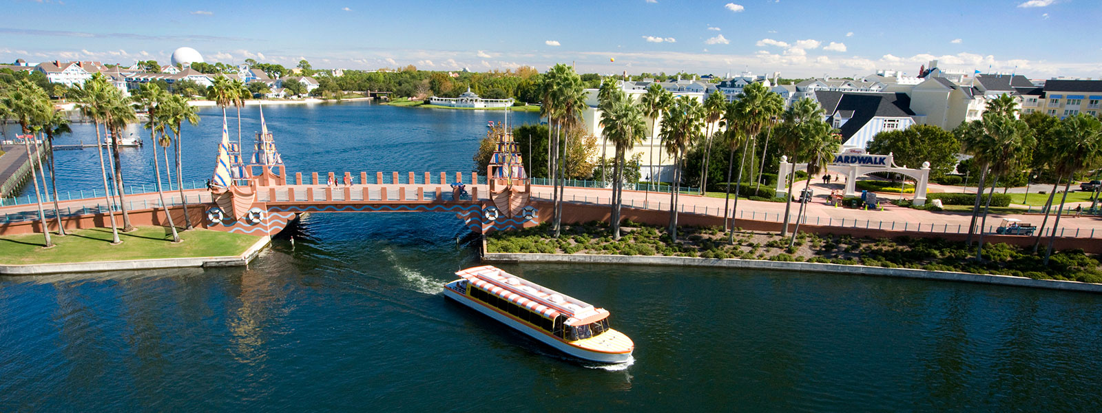 A boat travels along a canal under a decorative bridge, surrounded by palm trees and waterfront buildings with a sign for “Boardwalk” visible, all under a sunny blue sky.