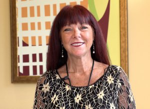 A woman with long red hair, wearing a black and beige floral patterned top, smiles at the camera. She has a headset and stands in front of a modern art painting with geometric shapes.