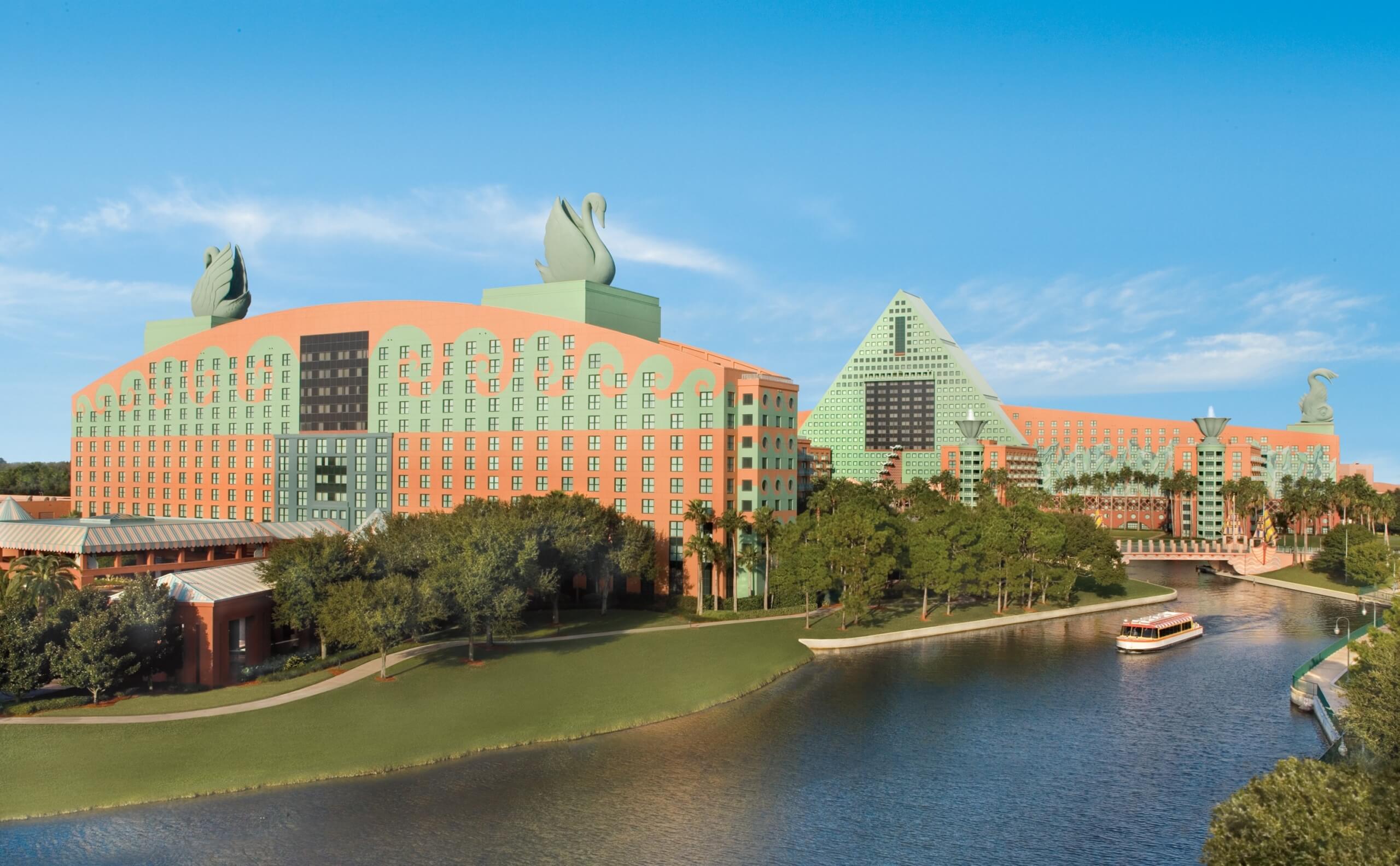Large pastel-colored hotel buildings with whimsical swan statues on the rooftops, next to a waterway with a boat, surrounded by trees under a clear blue sky.