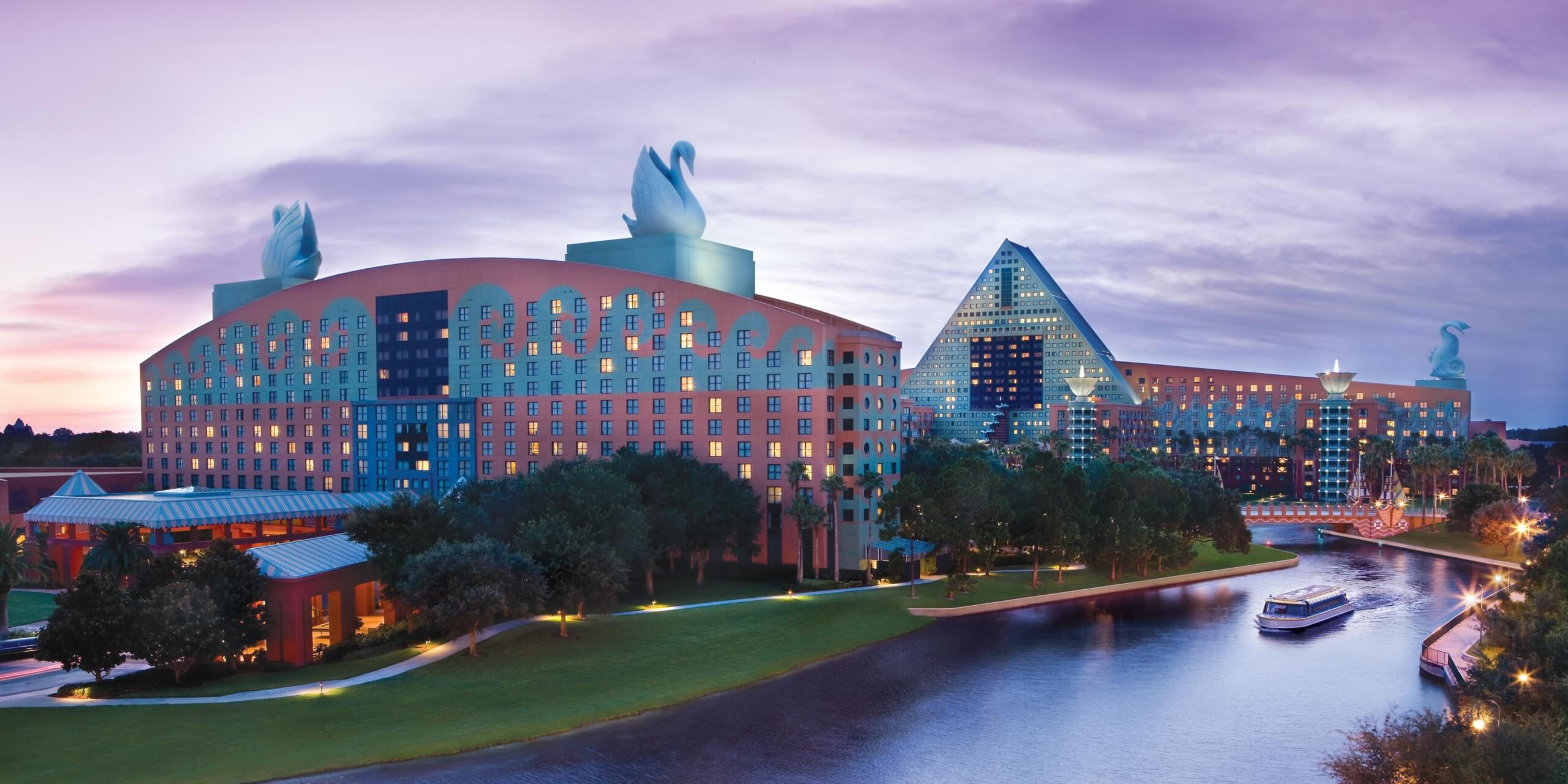 A large, lakeside hotel complex at sunset features a red and teal building with giant swan statues on the roof, a blue glass pyramid, lush landscaping, and a boat cruising on the water.