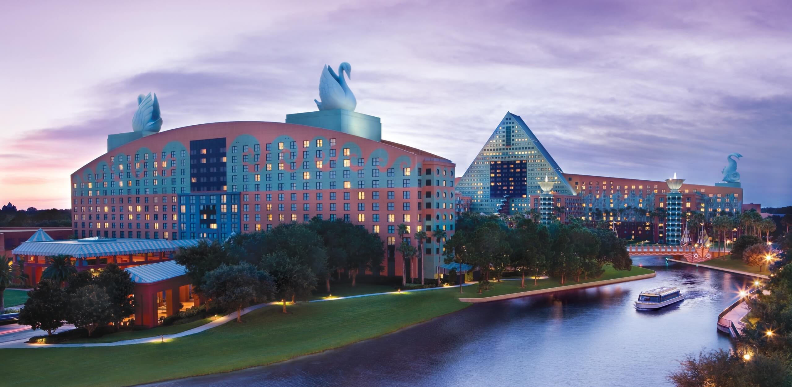 A large hotel complex with unique rooftop sculptures, surrounded by water and greenery, at sunset. A boat is traveling on the water near the buildings, and the sky is lit with soft purple and blue tones.