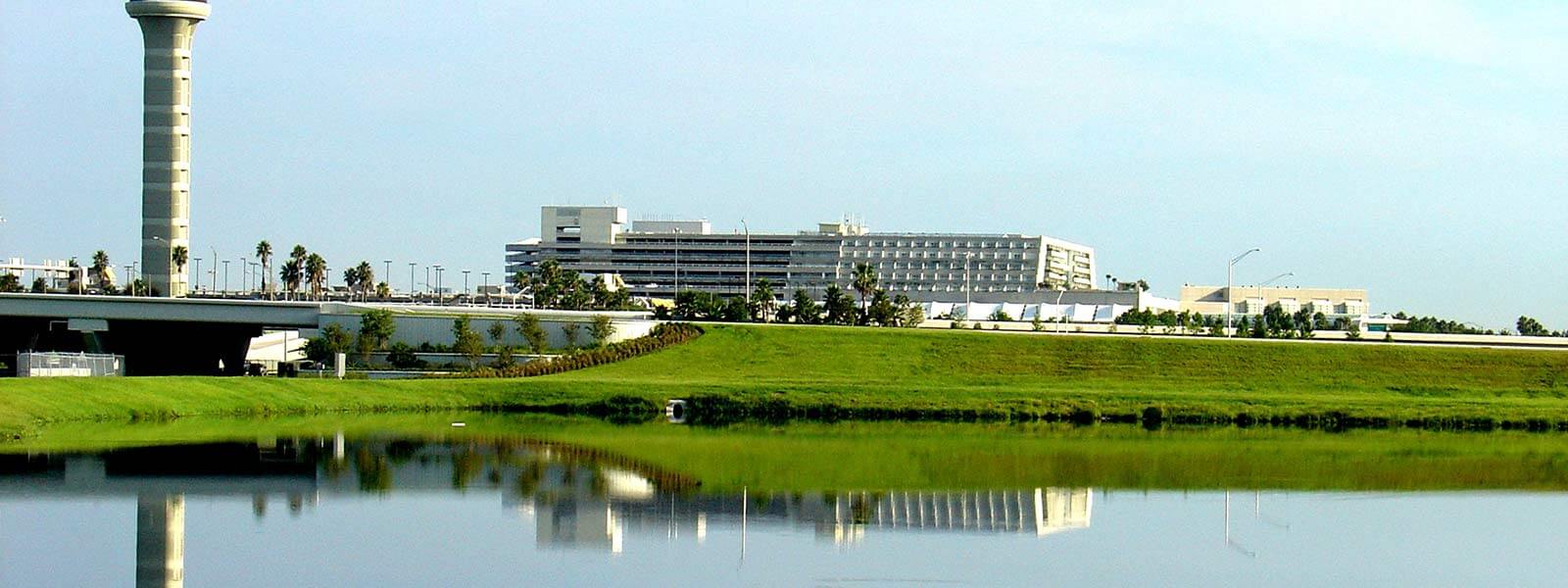 A modern airport control tower and terminal buildings are reflected in a calm body of water, surrounded by green grass and trees under a clear blue sky.
