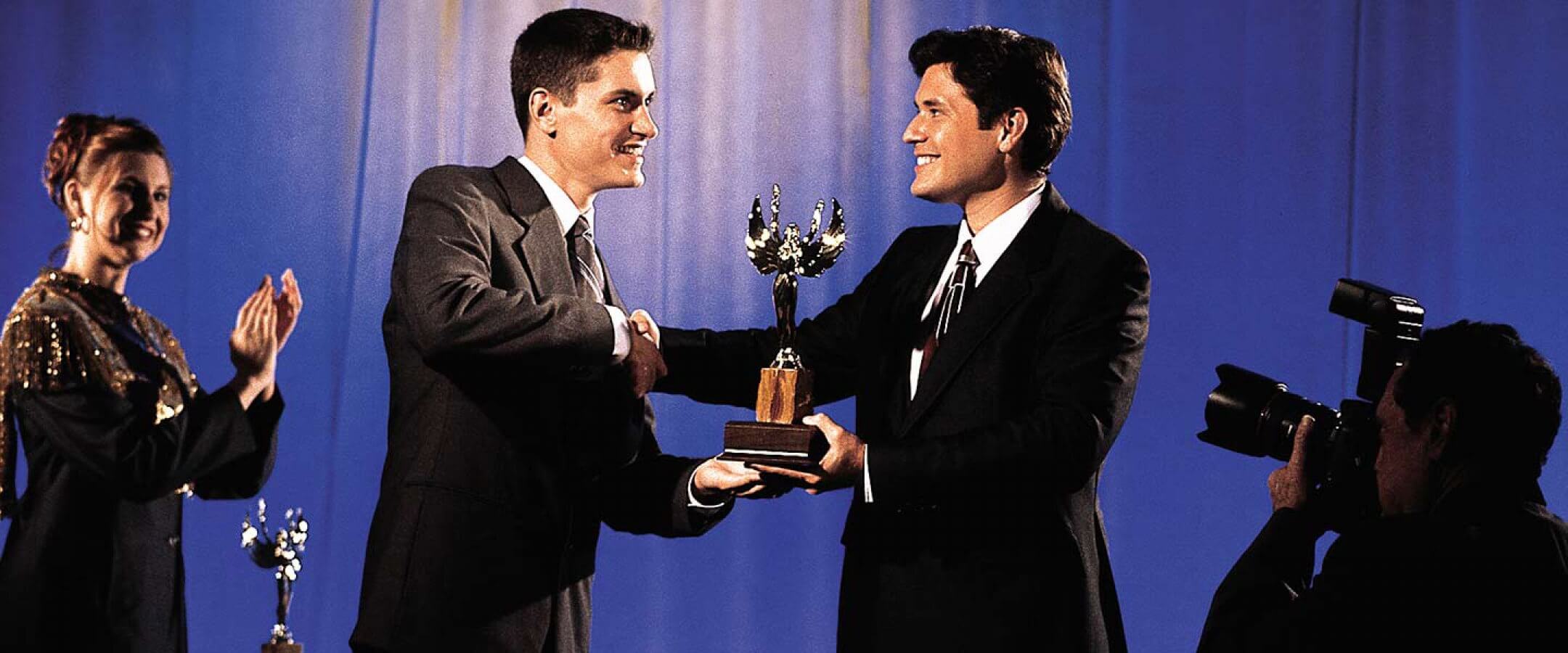 Two men in suits shake hands while one receives a trophy on stage; a woman claps in the background and a cameraman films the moment.