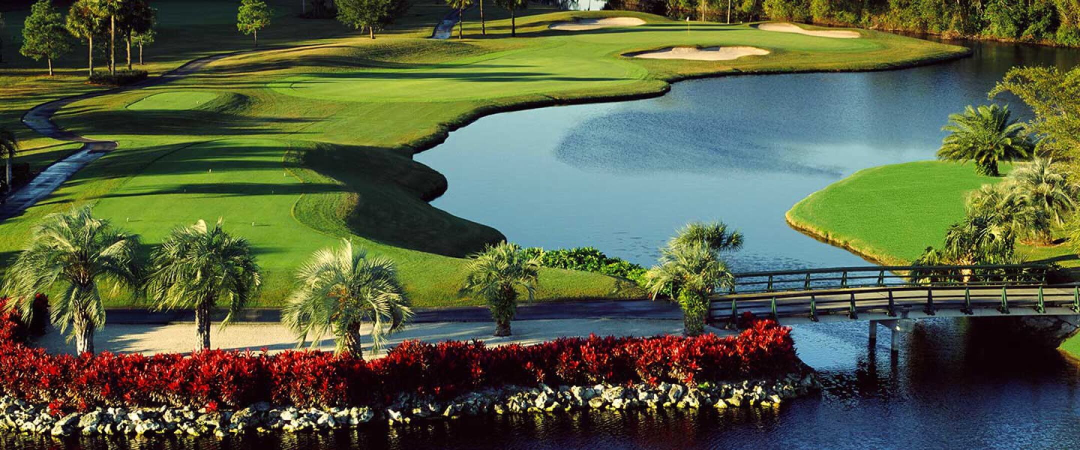 A scenic golf course featuring a lush green fairway, manicured sand bunkers, a tranquil pond, palm trees, red flowering shrubs, and a wooden bridge over the water under a clear sky.
