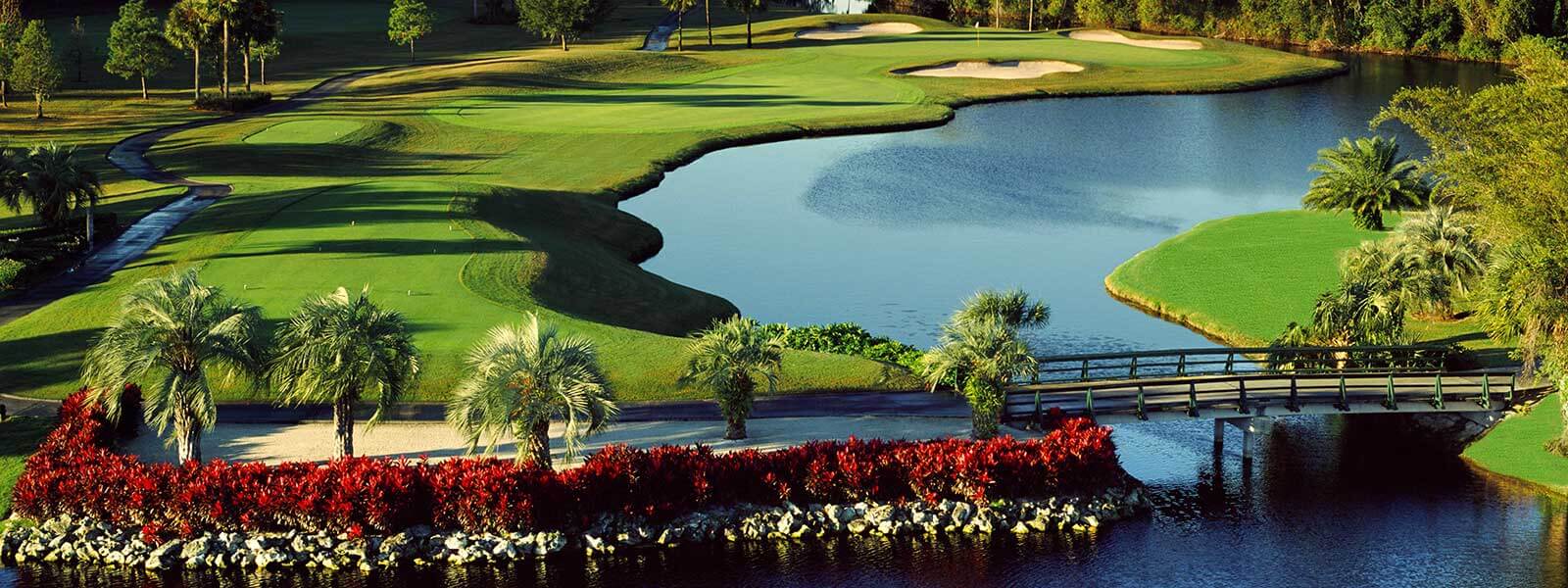A scenic golf course featuring a lush green fairway, manicured sand bunkers, a tranquil pond, palm trees, red flowering shrubs, and a wooden bridge over the water under a clear sky.