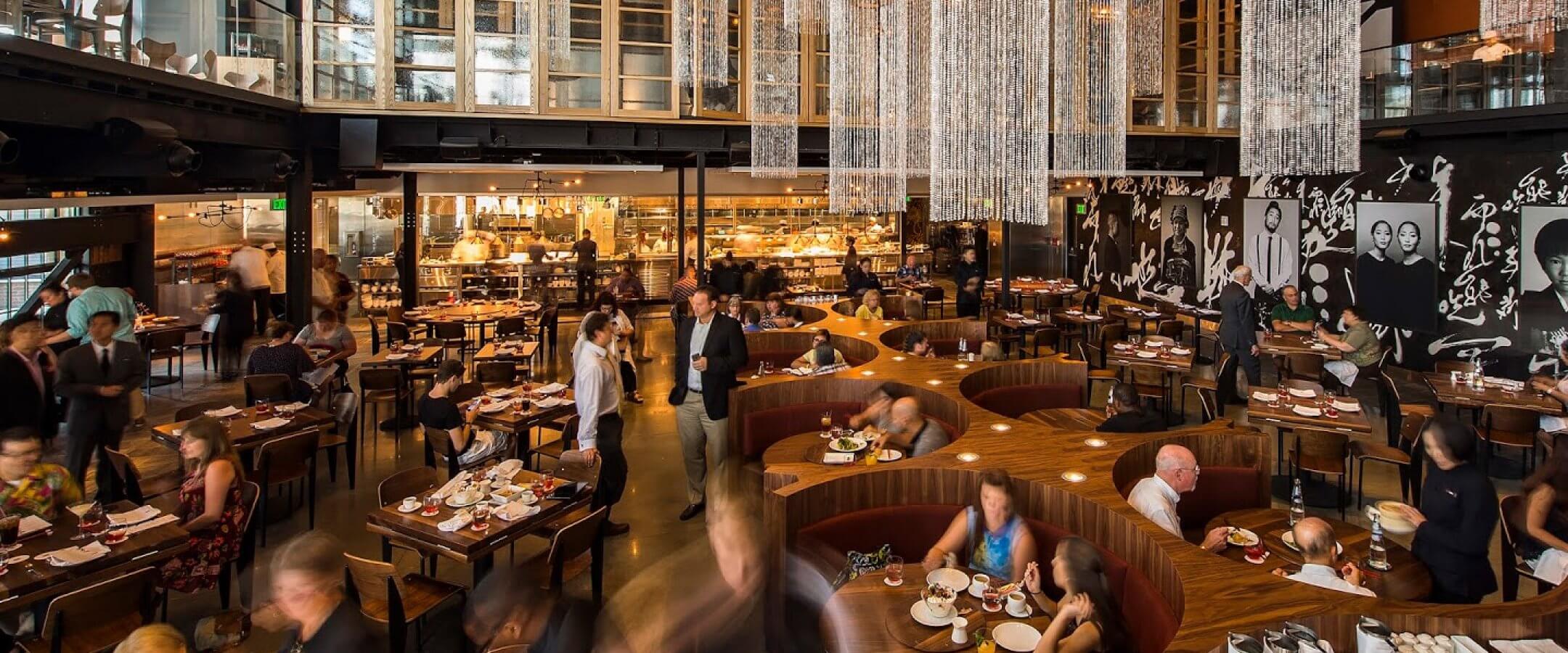 A stylish, modern restaurant with curved wooden seating, chandeliers, diners enjoying meals, an open kitchen in the background, and black-and-white portraits on the wall. The atmosphere is busy and lively.