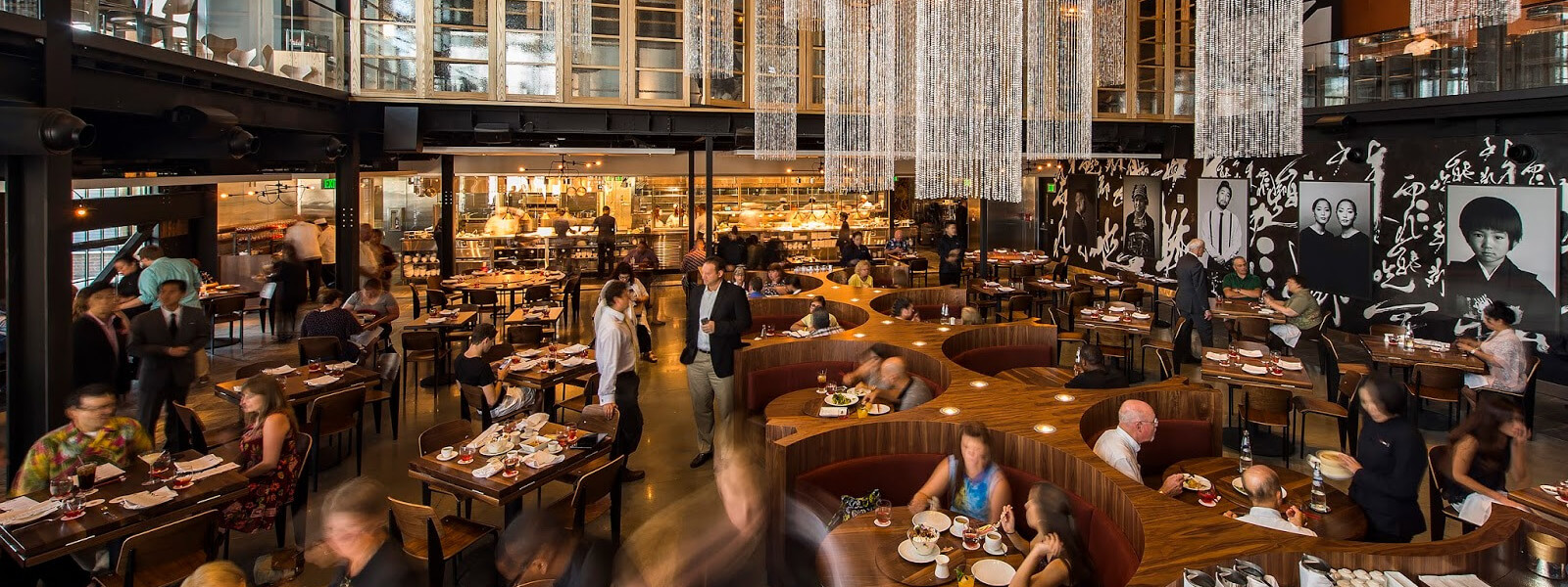 A stylish, modern restaurant with curved wooden seating, chandeliers, diners enjoying meals, an open kitchen in the background, and black-and-white portraits on the wall. The atmosphere is busy and lively.