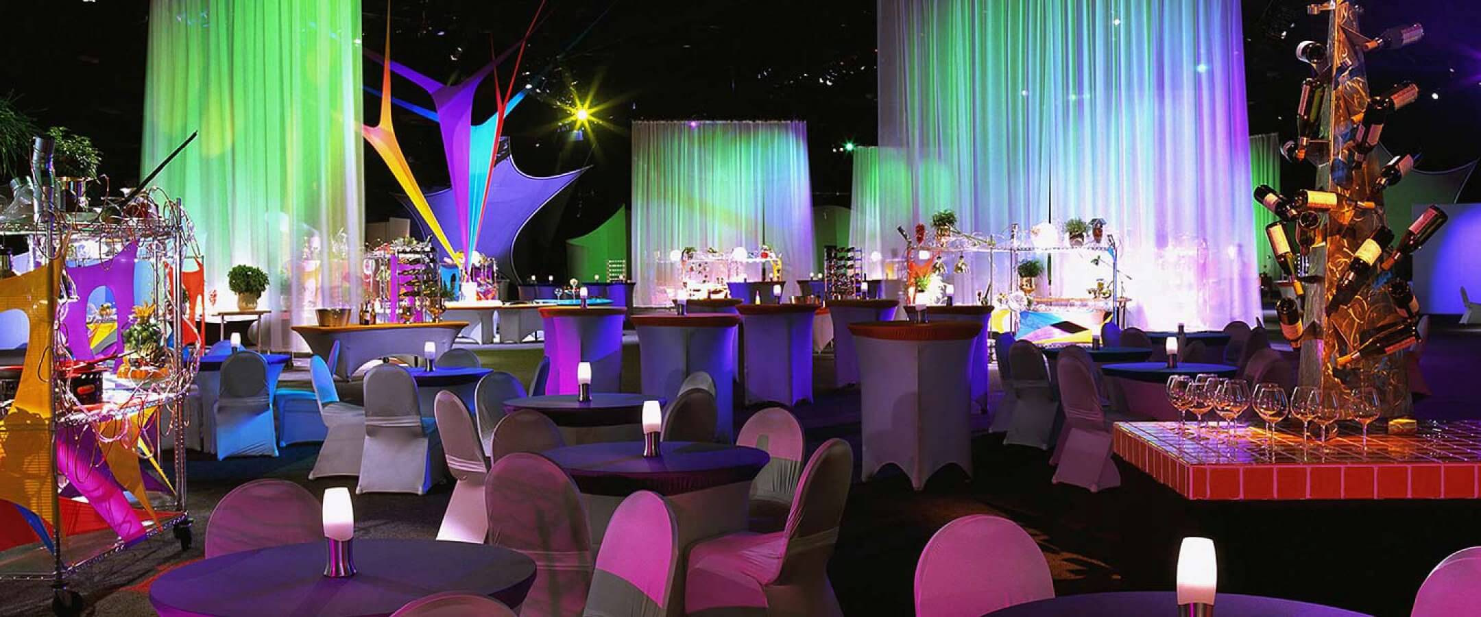 A colorful, modern event space with round tables and chairs draped in fabric, vibrant lighting, hanging curtains, decorative displays, and a wine bottle centerpiece, creating a festive, upscale atmosphere.