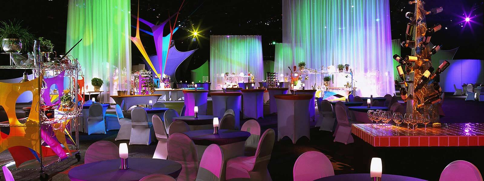 A colorful, modern event space with round tables and chairs draped in fabric, vibrant lighting, hanging curtains, decorative displays, and a wine bottle centerpiece, creating a festive, upscale atmosphere.