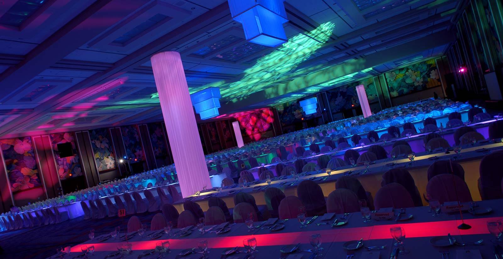 A large banquet hall with long tables set for dining, illuminated by colorful, vibrant lighting in shades of purple, blue, pink, and green, creating a festive atmosphere.