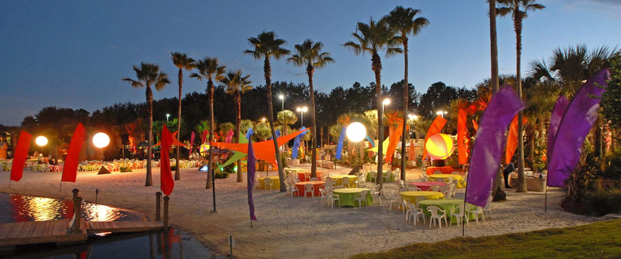 A festive outdoor beach party scene at dusk with colorful tables, chairs, and vibrant fabric banners among palm trees. Glowing round lanterns light up the sandy area near a small dock by the water.