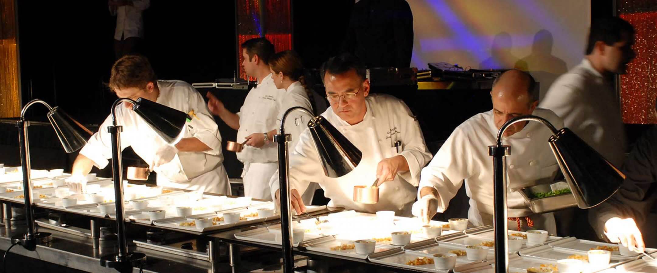 A group of chefs wearing white uniforms carefully plate dishes under bright lamps on a long table, preparing food in a busy, professional kitchen setting.