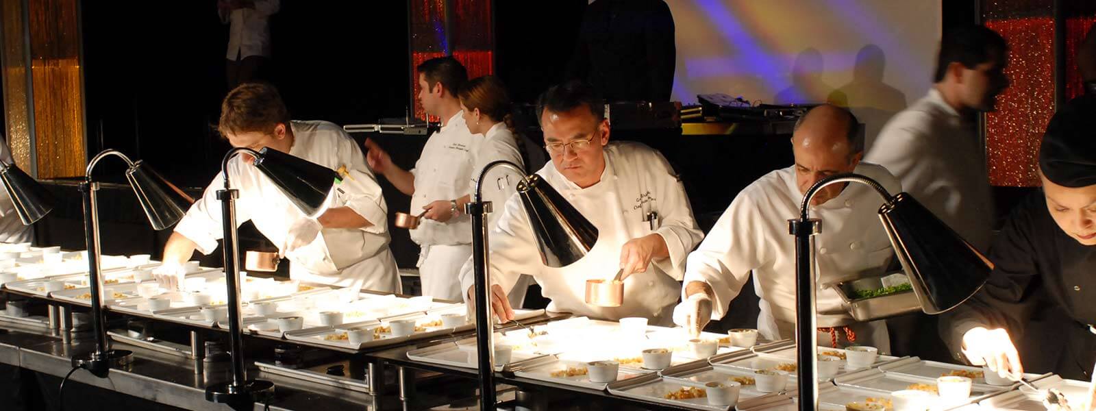 A group of chefs wearing white uniforms carefully plate dishes under bright lamps on a long table, preparing food in a busy, professional kitchen setting.