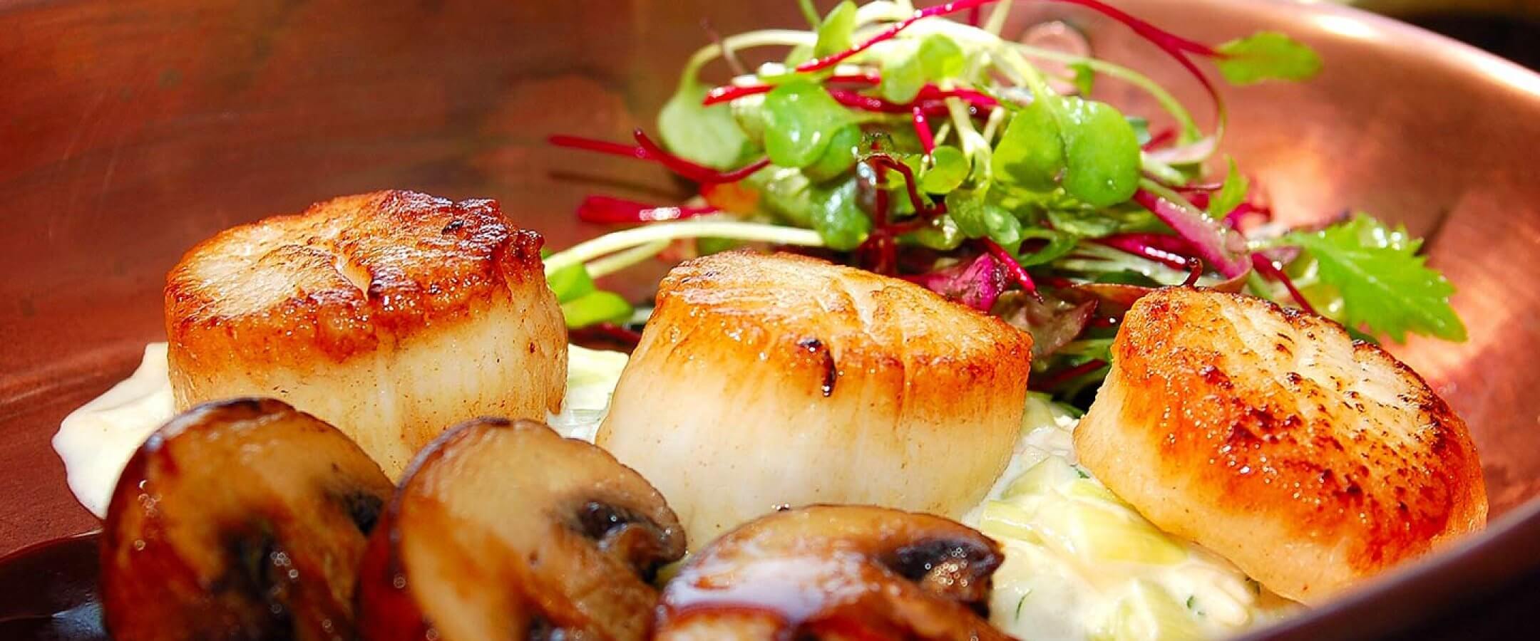 Three seared scallops served with sautéed mushrooms, creamy sauce, and a fresh mixed green salad on a copper plate.