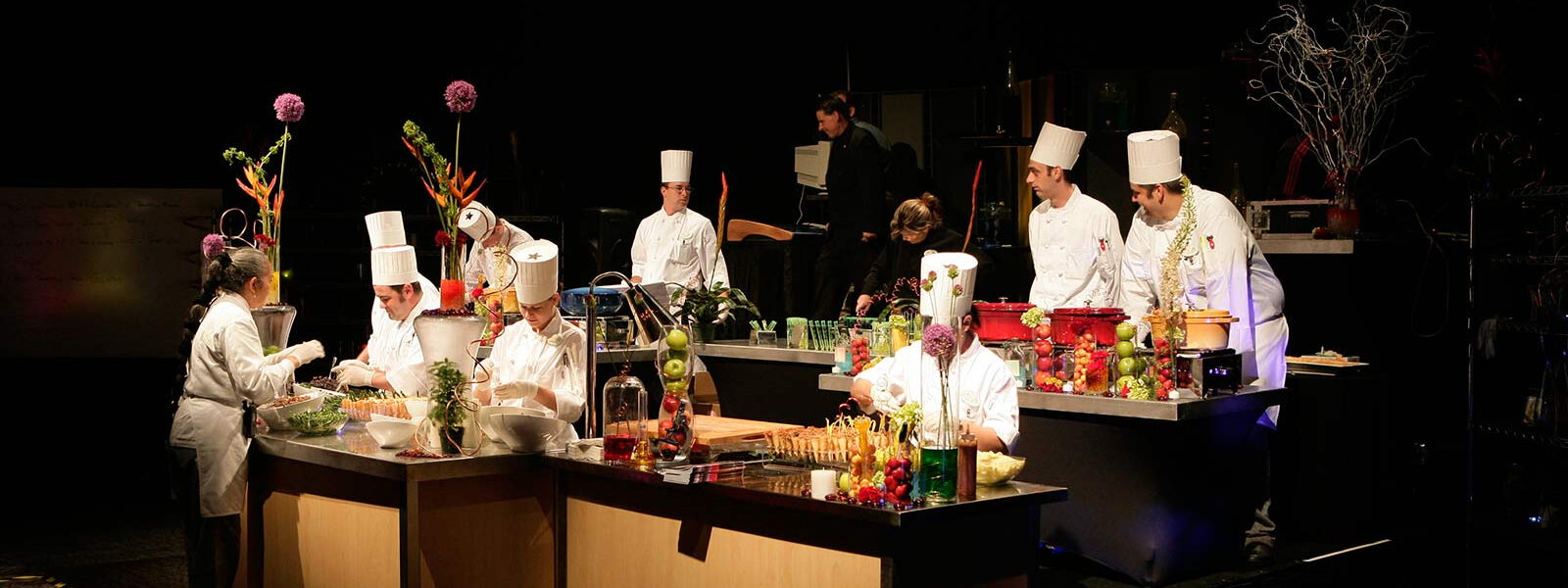 A group of chefs in white uniforms and hats prepare food at kitchen stations, surrounded by fresh ingredients and colorful garnishes, with people watching in the background under stage lighting.